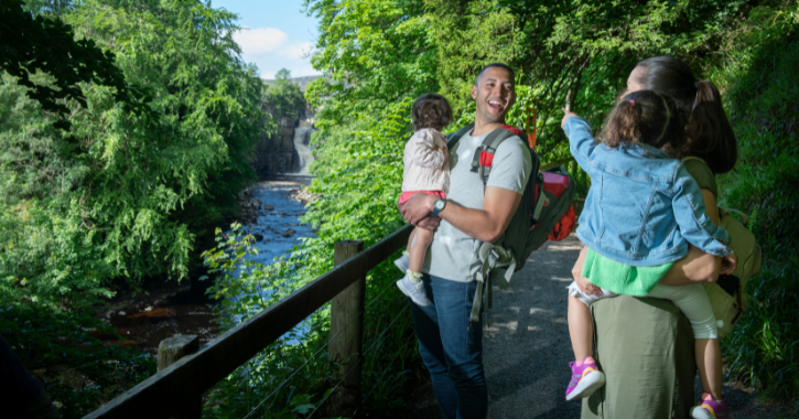 Man and woman standing smiling at each other while holding small children with High Force Waterfall in the background on a bright sunny day.